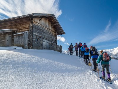 Line of people snowshoeing up snow-covered slope towards wooden hut on sunny day St Zyprian, Italy