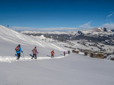 Group of people snowshoeing descending snow-covered hill following trail towards wooden huts with ski lift and mountains far into the distance, St Zyprian, Italy
