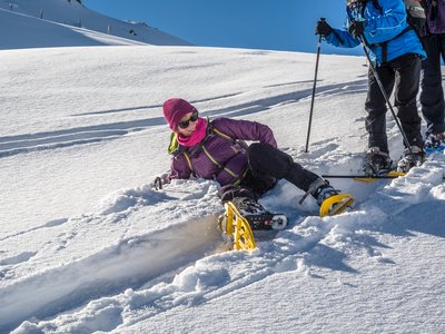 Lady smiling while lying on soft snow with snowshoe equipment on after having fallen during descent of snow-covered hill with people standing next to her, St Zyprian, Italy