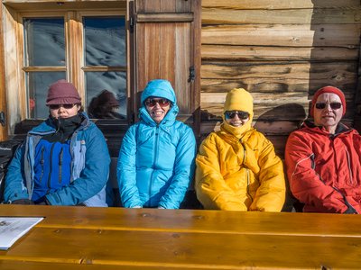 Walking group wearing colourful winter jackets sat outdoors at wooden table in front of wooden chalet with sun shining on them, St Zyprian, Italy