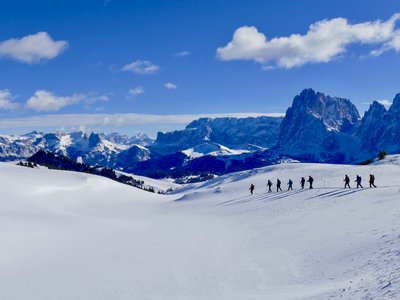 Snowy winter mountainous landscape with line of snowshoe walkers passing through with their long shadows being casted along the hill they descend, St Zyprian, Dolomites, Italy